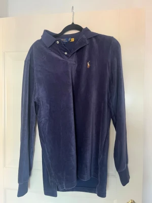 Blå långärmad skjorta Polo Ralph Lauren - Mörkblå långärmad skjorta från Polo Ralph Lauren i klassisk passform. Skjortan har mjukt sammetstyg, krage och knappar vid halsen samt den ikoniska broderade loggan på bröstet. Perfekt för dig som gillar stilrena och bekväma plagg.