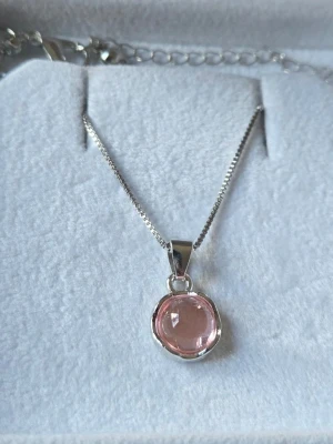 Silverhalsband med rosa sten - Snyggt halsband i silverfärg med en tunn kedja och ett runt hänge med en ljusrosa, glansig sten infattad i metall. Stenen har en mjuk, pärlliknande yta som ger ett elegant intryck. Perfekt accessoar för att lyfta din outfit. 45 cm + 5 cm förlängningskedja.