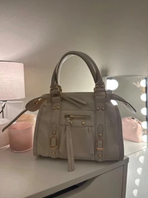 Beige handväska med gulddetaljer - Stilren beige handväska i skinnimitation med guldfärgade metalldetaljer och dragkedja framtill. Väskan har dubbla handtag och dekorativa sömmar samt en liten ficka med tofs. Perfekt accessoar för en trendig look. Använd en gång så skulle säga att den är som ny💖