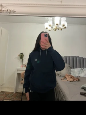 Mörkblå hoodie med logga - Mörkblå hoodie med huva och dragsnören, samt en liten vit logga på bröstet. Hoodien har en klassisk känguruficka framtill och ribbade muddar vid ärmslut och nederkant. Perfekt för en avslappnad och chill stil.