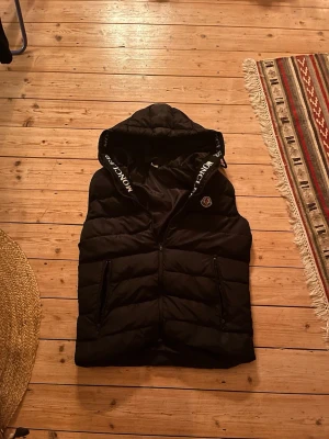 Svart dunväst från Moncler med huva - Snygg svart dunväst från Moncler med huva och dragkedja framtill. Västen har två fickor med dragkedja och Moncler-logga på bröstet. Huva med tryckt Moncler-text på kanten. Perfekt för lager-på-lager och streetwear.