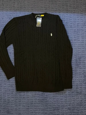 Svart kabelstickad tröja Polo Ralph Lauren - Kabelstickad svart tröja från Polo Ralph Lauren med rund halsringning och den klassiska vita broderade loggan på bröstet. Tröjan har långa ärmar och ribbade muddar. Perfekt för dig som gillar stilrena och tidlösa plagg.