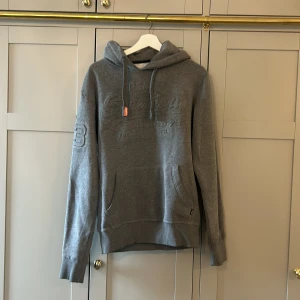 Grå hoodie från Superdry - Säljer en grå hoodie från Superdry med broderad logga på bröstet och fickor framtill. Tröjan har huva med dragsko och ribbade muddar vid ärmslut och nederkant. Perfekt för en avslappnad stil.