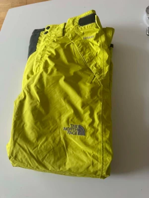 Gula skalbyxor från The North Face - Säljer ett par gula skalbyxor från The North Face med justerbar midja och dragkedja. Byxorna har förstärkta partier i svart vid bensluten och är tillverkade i slitstarkt, vattenavvisande material. Perfekta för skidåkning eller friluftsliv.