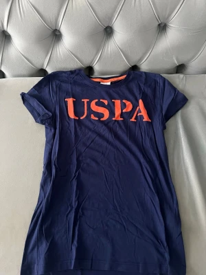 Mörkblå USPA t-shirt med orange tryck - Snygg mörkblå t-shirt från U.S. Polo Assn. med stort orange USPA-tryck på bröstet. T-shirten har rund hals med orange kant och är tillverkad i mjuk bomull. Perfekt för en avslappnad och sportig stil.