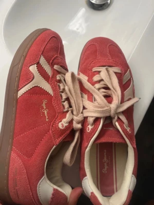 Röda/rosa sneakers från Pepe Jeans - Snygga rosa sneakers från Pepe Jeans i mocka med beige detaljer och matchande beige/ljusrosa snörning. Skorna har en låg siluett och gummisula. Använda 2 gånger så i väldigt bra skick