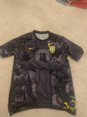 Svart Brasilien t-shirt Nike - Säljer en svart t-shirt från Nike med Brasilien-emblem och ett stort grafiskt tryck av Kristusstatyn på framsidan. T-shirten har korta ärmar, rund hals och detaljer i gult. Perfekt för dig som gillar unika fotbollströjor.