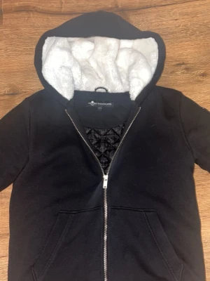 Svart hoodie från Moose Knuckles - Säljer en svart hoodie från Moose Knuckles med dragkedja och vit fluffig foder i huvan. Hoodien har fickor framtill och en ikonisk logga på bröstet med silverdetaljer. Perfekt för dig som vill ha både stil och komfort, storlek xl i barnstorlek men passar som xs.
