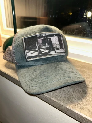 Grå meshkeps med tryck framtill - Säljer en grå keps med mesh baktill och justerbar snapback. Framtill finns ett svartvitt tygmärke med ett grafiskt motiv av en person som går över ett övergångsställe. Perfekt för dig som gillar streetstyle och unika detaljer.