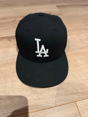 Svart LA-keps med rak skärm - Säljer en svart keps från New Era med klassisk LA-logga broderad i vitt framtill. Kepsen har rak skärm och ventilationshål upptill. Perfekt för dig som gillar streetwear och sportig stil. Skärmen har böjts lite av mig själv men inte Brutits, köpt på New Era storleken är tagen på skärmen är borttagen men kan real testas vid behov.