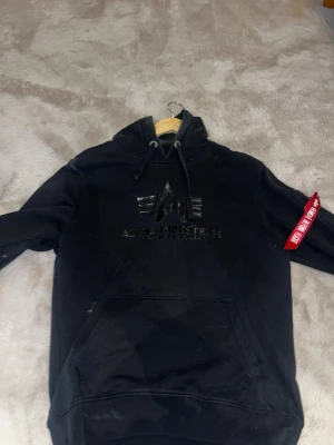 Svart hoodie från Alpha Industries - Säljer en svart hoodie från Alpha Industries med stor logga på bröstet och klassisk röd tag på ärmen. Hoodien har dragsko i huvan och en stor magficka. Tillverkad i mjukt bomullsmaterial och har ribbade muddar vid ärmslut och nederkant.