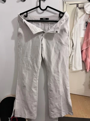 Vita kostymbyxor från Bikbok - Snygga vita kostymbyxor från Bikbok med bootcut ben och diskreta fickor bak. Byxorna har klassisk knapp- och dragkedjestängning samt är tillverkade i ett mjukt material som ger en clean look. Perfekta för dig som gillar stilrena och minimalistiska outfits. Byxorna har även en slits längst ner