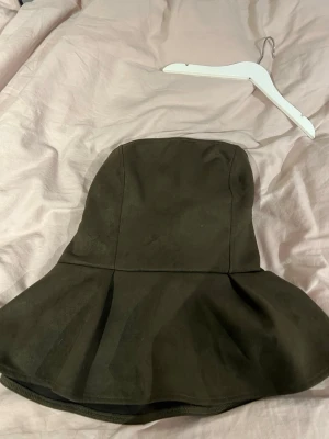 Mörkgrön tubtopp - Supersnygg mörkgrön tubtopp i mockaimitation med peplumskärning. Toppen är axelbandslös och har en figurnära passform upptill som sedan går ut i en volang nedtill. Perfekt för dig som vill ha en trendig och unik topp.