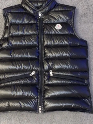 Svart dunväst från Moncler - Snygg svart dunväst från Moncler med glansig finish och klassisk logga på bröstet. Västen har två dragkedjeförsedda fickor och hög krage. Perfekt för lager-på-lager och riktigt trendig look. Fylld med dun för extra värme. Ny skick