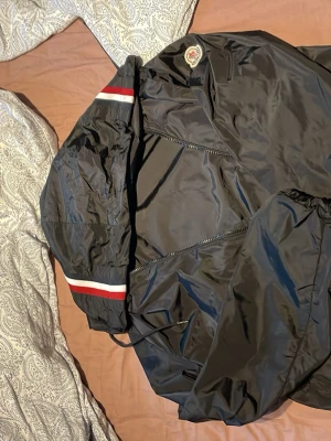 Svart vindjacka från Moncler - Snygg svart vindjacka från Moncler med huva och dragkedja. Jackan har Moncler-logga på bröstet och rödvit rand på huvan. Tillverkad i glansigt, lätt material som står emot vind. Perfekt för dig som vill ha en stilren och sportig look.