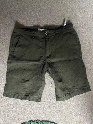 Olivgröna linneshorts - Nypris i butik: 499kr                           Snygga olivgröna shorts i linne från Dressman med normal passform. Shortsen har sidofickor fram, bälteshällor och knäppning med knapp och dragkedja. Baktill finns två fickor med knappstängning. Perfekta för varma dagar. Linnechinos.