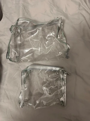 Två genomskinliga necessärer - Säljer två genomskinliga necessärer med silverfärgade detaljer och dragkedja. Perfekta för att organisera smink, småprylar eller researtiklar. Båda väskorna är i plastmaterial och har en stilren, enkel design.