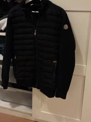 Svart pufferjacka från Moncler - Säljer en svart pufferjacka från Moncler med quiltad front, två dragkedjefickor och klassisk Moncler-logga på ärmen. Jackan har hög krage och dragkedja framtill. Materialet är en mix av dun och syntet, vilket ger både värme och stilren look.