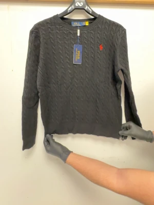 Mörkgrå kabelstickad tröja Polo Ralph Lauren - Snygg mörkgrå kabelstickad tröja från Polo Ralph Lauren med rund halsringning och röd broderad logga på bröstet. Tröjan har långa ärmar och ribbade muddar. Perfekt för dig som gillar klassisk och stilren design.