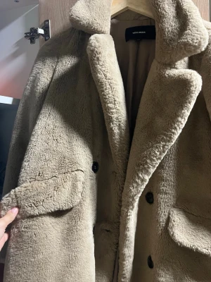 Beige teddyjacka / kappa  från Vero Moda - Säljer en supermjuk och fluffig teddyjacka från Vero Moda i beige färg. Jackan har stora slag, knäppning framtill med svarta knappar och fickor med lock. Perfekt för dig som vill ha en trendig och mysig look under kyliga dagar. Priset går att diskutera