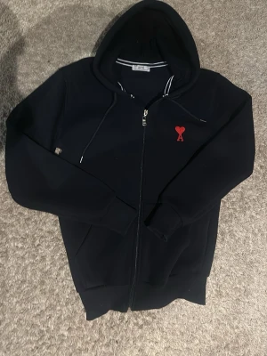  hoodie från Ami Paris - Snygg svart hoodie från Ami Paris med dragkedja och huva. På bröstet sitter Ami:s ikoniska röda hjärta med A. Tillverkad i mjukt material och har dragsko i huvan samt fickor framtill. Perfekt för en chill och stilren look.