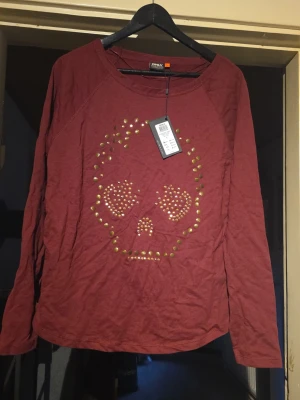 Röd långärmad topp med döskalle - Cool röd långärmad topp från Only med guldfärgade nitar som bildar ett döskallemotiv på framsidan. Rund halsringning och avslappnad passform. Perfekt för dig som vill sticka ut med en edgy detalj i din outfit.
