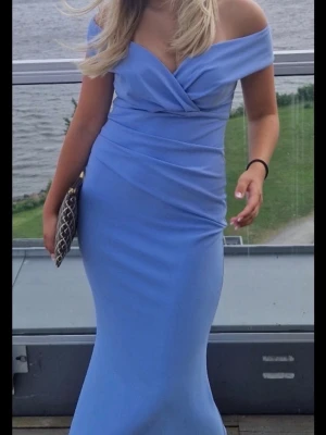 Ljusblå långklänning med omlott - Elegant ljusblå långklänning med omlottdetalj upptill och drapering i midjan. Klänningen har v-ringad hals och är ärmlös, vilket ger en stilren och modern look. Perfekt för speciella tillfällen där du vill känna dig extra fin som t.ex på balen.