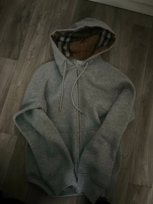 Grå zip hoodie från Burberry - Snygg grå zip hoodie från Burberry med klassiskt rutigt foder i huvan och dragsko. Tillverkad i mjuk bomullsblandning och har ribbade muddar samt fickor framtill. Perfekt för en avslappnad och trendig look.
