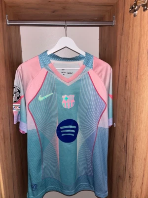 Barcelona matchtröja Lamine Yamal - Säljer en Barcelona matchtröja från Nike i ljusblått och rosa med tryck av Lamine Yamal och nummer 10 på ryggen. Tröjan har officiella klubbmärket, Nike-logga och Spotify sponsortryck på bröstet. Tillverkad i lätt och ventilerande material, perfekt för fotboll.