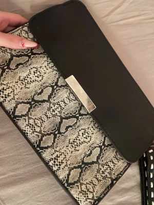 Svart kuvertväska med ormmönster - Snygg kuvertväska i svart konstläder med lock och detaljer i silverfärgad metall. Framsidan har ett trendigt ormmönster i svart och beige som ger väskan en edgy look. Perfekt att bära i handen för en stilren outfit. Material av skinn