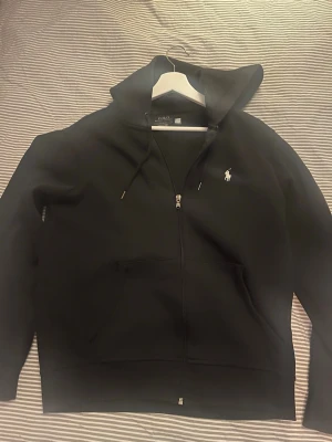 Svart zip hoodie från Polo Ralph Lauren - Snygg svart hoodie med dragkedja från Polo Ralph Lauren. Klassisk design med vit broderad logga på bröstet, huva med dragsko och två fickor framtill. Perfekt för en avslappnad och stilren look.