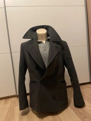 Tiger of sweden rock - Stilren mörkgrå skepparkavaj med dubbelknäppning och bred krage. Jackan har två framfickor och en bröstficka, samt klassisk figursydd passform. Tillverkad i ett tjockare tyg som passar perfekt för kyligare dagar. Inga synliga loggor eller märken.