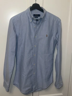 Ljusblå skjorta från Polo Ralph Lauren - Klassisk ljusblå skjorta från Polo Ralph Lauren med button-down krage och broderad logga på bröstet. Skjortan har långa ärmar, knappar framtill och är tillverkad i mjuk bomull. Perfekt för en clean och stilren look.