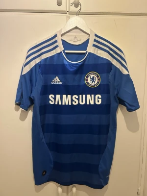 Chelsea FC 11/12  Drogba #11 - Chelsea FC matchtröja från Adidas i blått med vita detaljer och ränder på axlarna. Tryck med Drogba #11 på ryggen och Samsung-logga på bröstet. Kortärmad och tillverkad i lätt polyester, perfekt för fotbollsträning eller match.