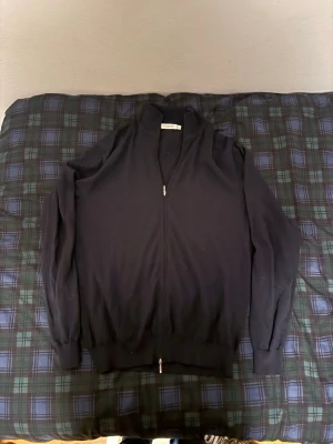 Mörkblå merinoullströja från Suitsupply - Snygg mörkblå stickad tröja från Suitsupply i mjuk merinoull. Tröjan har dragkedja framtill, hög krage och långa ärmar. Perfekt för dig som vill ha en stilren och bekväm look med premiumkänsla. Liten i storleken passar mer som M