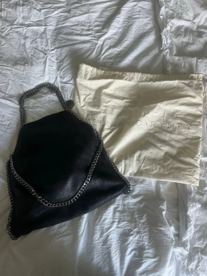 Svart Stella McCartney Falabella väska - Säljer en svart Stella McCartney Falabella handväska i veganskt mockaliknande material. Väskan har silverfärgade kedjedjor som ej rostat. Väskan är rymlig och du får plats med en 15tums dator i. Måtten är 37x36x9. Perfekt som vardagsväska eller till festligare tillfällen. Innertyget är lite smutsigt men annars är väskan i fint skick. Dustbag tillkommer. Tveka inte på att skriva om ni har ytterligare frågor eller prisförslag❤️
