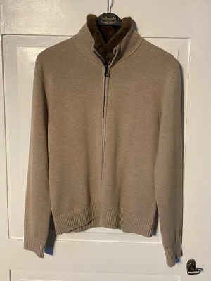 Beige cardigan kofta med avtagbar pälskrage - En beige kofta med dragkedja i fullängd samt avtagbar pälskrage. Ett häftigt plagg för dig som vill ha mångsidighet. Inga lappar eller etiketter på tröjan som indikerar storlek men uppskattat den till medium. Mycket bra skick, inget tecken på användning vare sig på själva tröjan eller på kragen. Bara att skriva vid funderingar