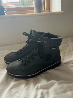 Svarta kängor från Rieker - Säljer ett par svarta kängor från Rieker med snörning och grov sula. Skorna har en stickad grå mudd upptill och detaljer i rött på snörena. Tillverkade i syntetmaterial med dekorativa sömmar och rund tå. Använda enstaka gånger, så i princip nyskick