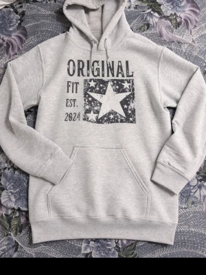 Grå hoodie med stjärntryck - Snygg ljusgrå hoodie med stor ficka framtill och tryck med texten 'ORIGINAL FIT EST. 2024' samt en stor stjärna. Hoodien har huva med dragsko och ribbade muddar vid ärmar och nederkant. Perfekt för en avslappnad streetwear-look.