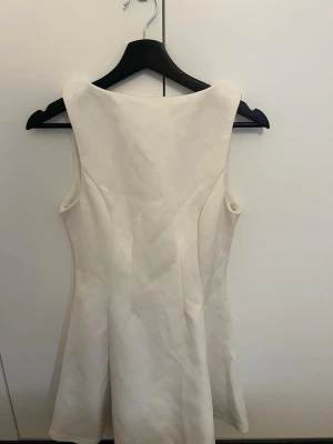 Vit ärmlös kortklänning - Säljer en stilren vit kortklänning med ärmlös design och rund hals. Klänningen har en lätt utsvängd form och dragkedja i ryggen. Perfekt för dig som gillar enkel och klassisk stil.