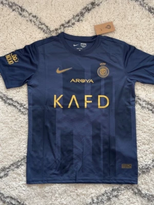 Al Nassr Ronaldo matchtröja blå - Snygg blå matchtröja från Al Nassr med guldiga detaljer och tryck. Framsidan har klubbmärke, Nike-logga och sponsortryck i guld. Baksidan har namnet 'Ronaldo' och nummer 7 i guld. Tillverkad i lätt och ventilerande funktionsmaterial, perfekt för fotbollsträning.