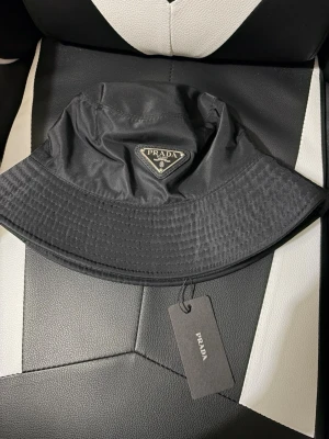 Svart bucket hat - Stilren svart bucket hat från Prada med klassisk logga framtill i metall och tyg. Hatten är tillverkad i nylon och har en mjuk, rundad form med bred skärm. Perfekt accessoar för en trendig streetwear-look.