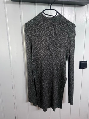 Mörkgrå ribbad långärmad klänning/tunica - Mysig mörkgrå ribbad klänning/tunica med lång ärm och hög hals. Klänningen har en slits på sidan för extra rörelsefrihet och är i ett mjukt stickat material. Perfekt för dig som gillar enkel men stilren design.