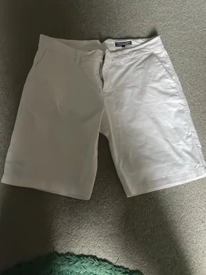 Vita shorts från Tommy Hilfiger - Säljer ett par klassiska vita shorts från Tommy Hilfiger med normal passform. De har fickor fram och bak, bälteshällor och stängs med dragkedja och knapp. Perfekta till sommaren och lätta att matcha med olika stilar. 