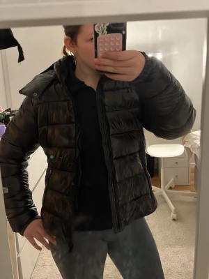 Svart pufferjacka med huva - Säljer en svart pufferjacka med huva och dragkedja framtill. Jackan har en quiltad design med breda kanaler och är perfekt för kalla vinterdagar. Den har en klassisk passform och är lätt att matcha till olika outfits. ALDRIG använd, lapp kvar 