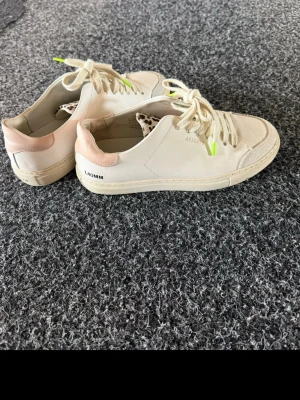 Beige sneakers från ARIGATO - Snygga beige sneakers från ARIGATO med ljusrosa detalj på hälen och vita snören med limegröna detaljer. Skorna har en klassisk låg profil och är tillverkade i slätt läder. Perfekta för dig som gillar stilrena och trendiga sneakers.