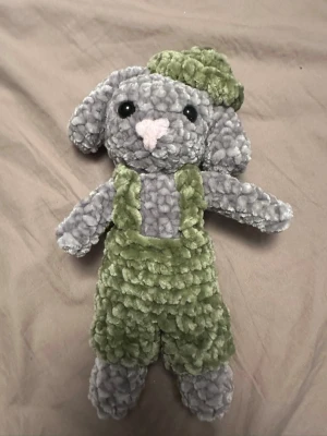 Grå mjuk kanin i gröna kläder - Supermjuk grå kanin med säkerhets ögon och gröna hängselbyxor samt matchande grön mössa. Kaninen har långa öron och en liten svans bak. Perfekt som gosedjur eller dekoration. Är ca 20-25 cm 