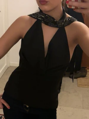 Svart halterneck topp med paljettdetaljer - Snygg svart halterneck topp med draperad front och breda band runt halsen dekorerade med svarta paljetter. Toppen är ärmlös och har en croppad passform som ger en modern och trendig look. Perfekt för dig som vill sticka ut.