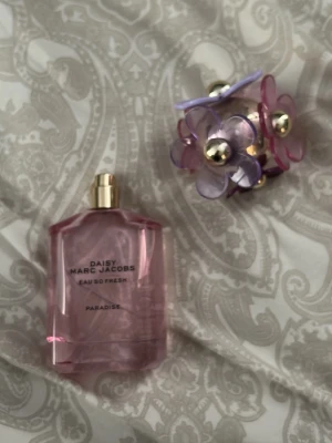 Daisy Eau So Fresh Paradise parfym - Daisy Eau So Fresh Paradise från Marc Jacobs är en parfym i en genomskinlig rosa flaska med guldfärgad spraykork. Locket är dekorerat med stora plastblommor i lila och rosa med gulddetaljer, vilket ger en lekfull och färgstark look. Nästan allt kvar skulle gissa runt 70 ml