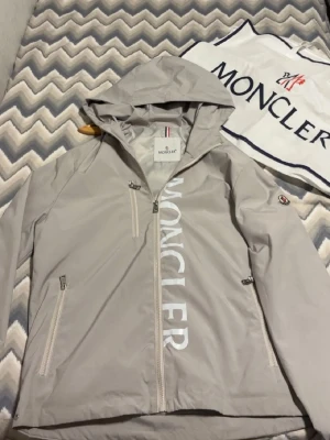 Grå vindjacka från Moncler med huva - En helt ny moncler jacka. Jag säljer den då den är för liten för mig. Den passar en liten M                                                                                Snygg grå vindjacka från Moncler med stor logga framtill, huva med dragsko och dragkedjor på fickorna. Jackan har långa ärmar, diskret Moncler-märke på ärmen och är tillverkad i lätt polyester. Perfekt för dig som gillar stilren och sportig look.